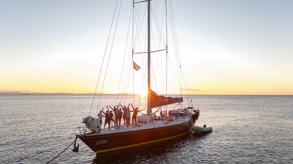 Wide shot of a sailing boat on the Whitsundays during sunset with a group of backpackers on deck.