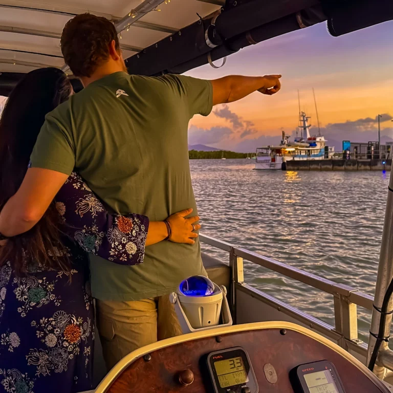 A man and a woman holding onto each other whilst pointing out something in the distance during a sunset river cruise in Cairns.