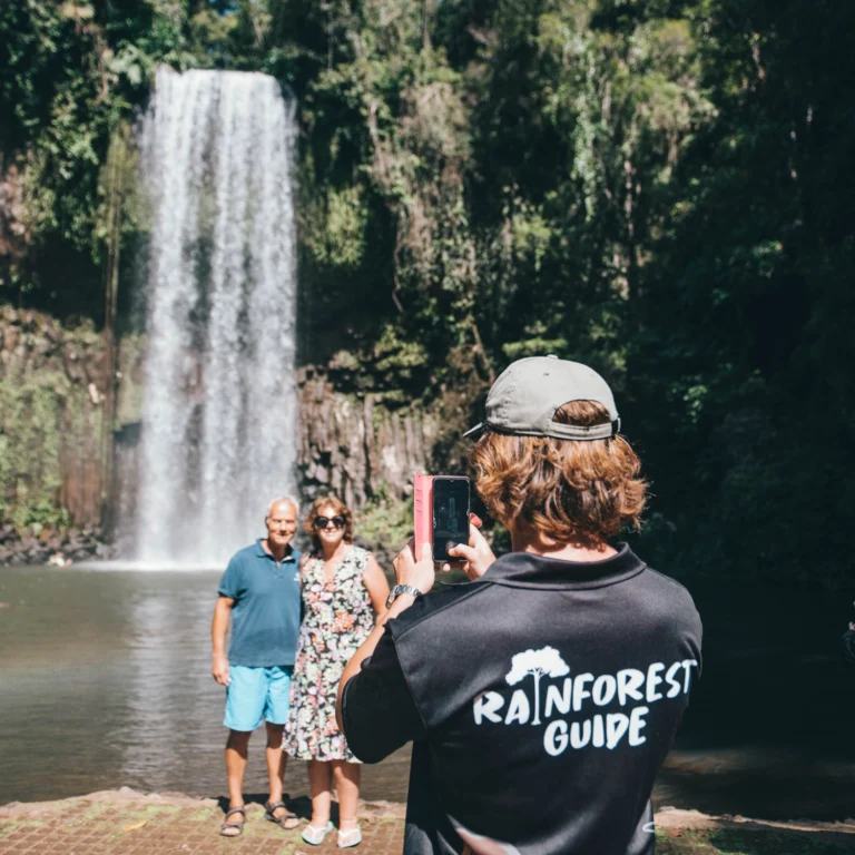 A rainforest guide has his back to the camera. He is taking a photo of a couple visting the waterfalls of the Atherton Tablelands