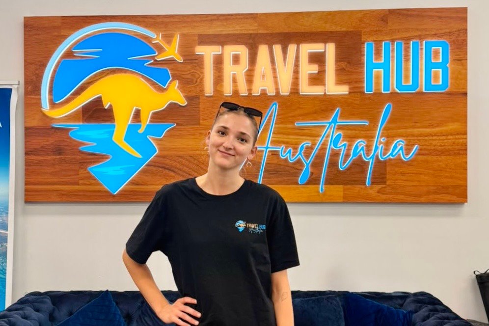 A photo of Elyra, a Travel Hub Specialist standing infront of the Travel Hub logo
